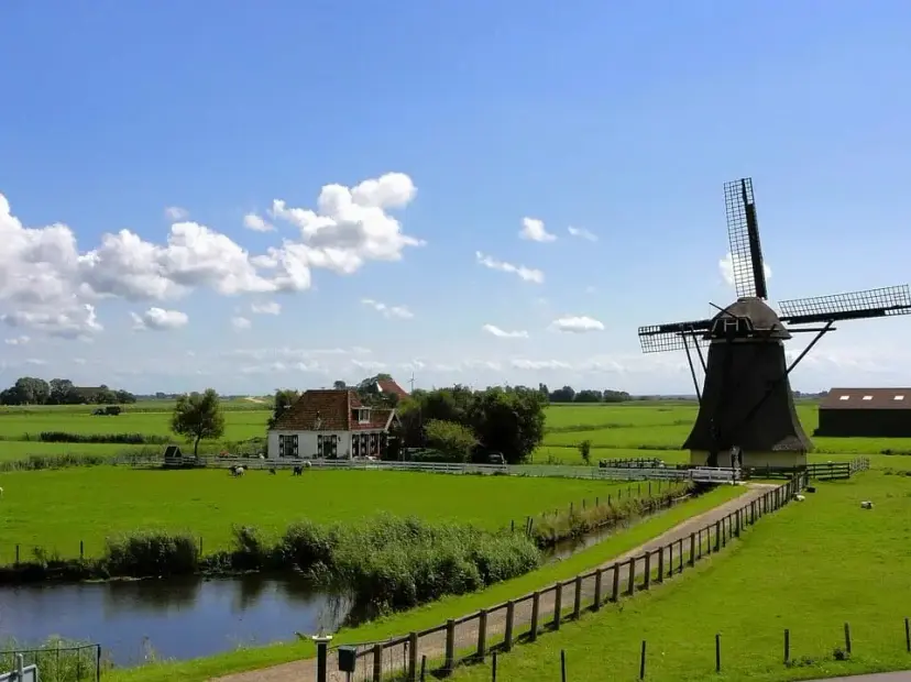 Dutch windmill beside green pastures, a farmhouse and a canal under a blue sky with clouds.