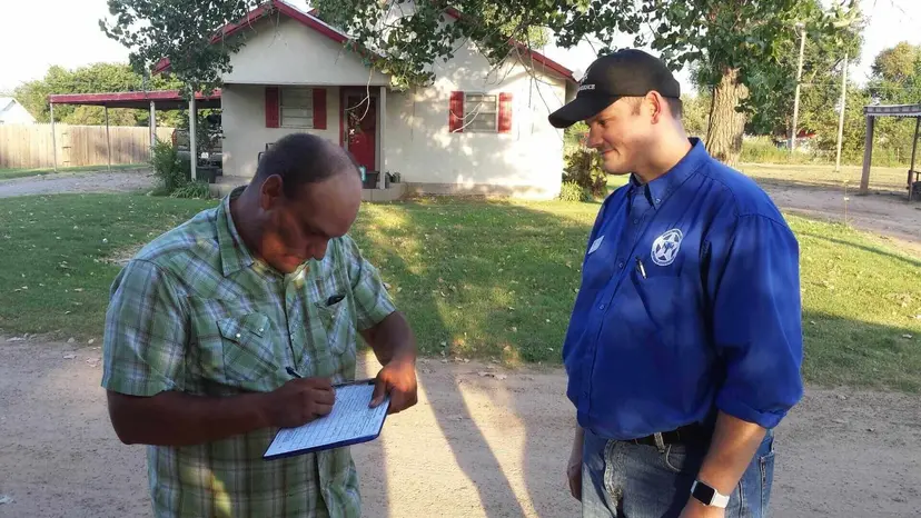 A man signs a clipboard while a man in a blue TNM shirt stands beside him outside a house