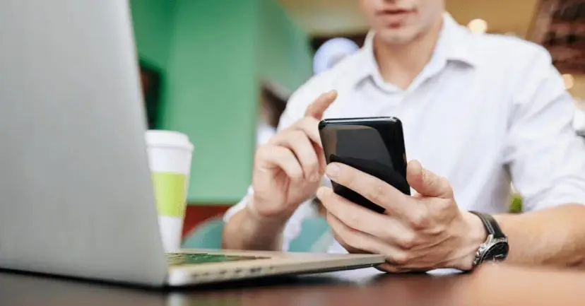 A man checks his smartphone at a table beside an open laptop and a coffee cup.