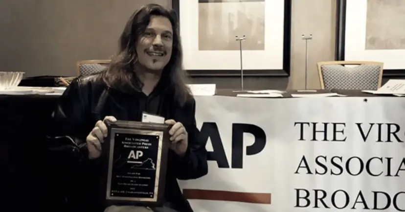 A long-haired man smiles while holding a Virginia Associated Press Broadcasters award plaque beside an AP banner.