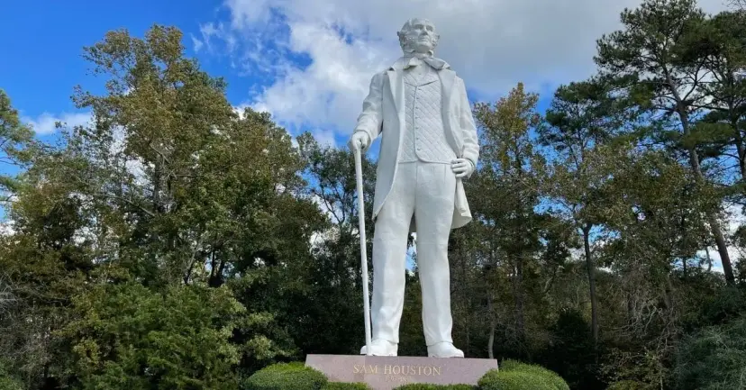 A tall white statue of Sam Houston holding a cane, surrounded by trees.