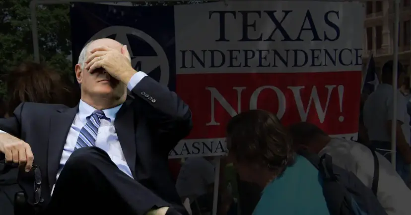 A man in a suit covers his face with his hand in front of a 'Texas Independence Now!' banner.