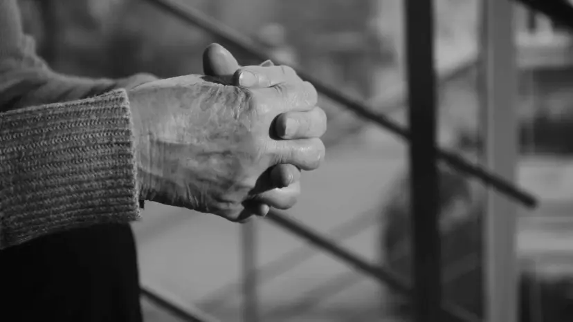 Black and white photo of an elderly person's clasped hands resting on a railing