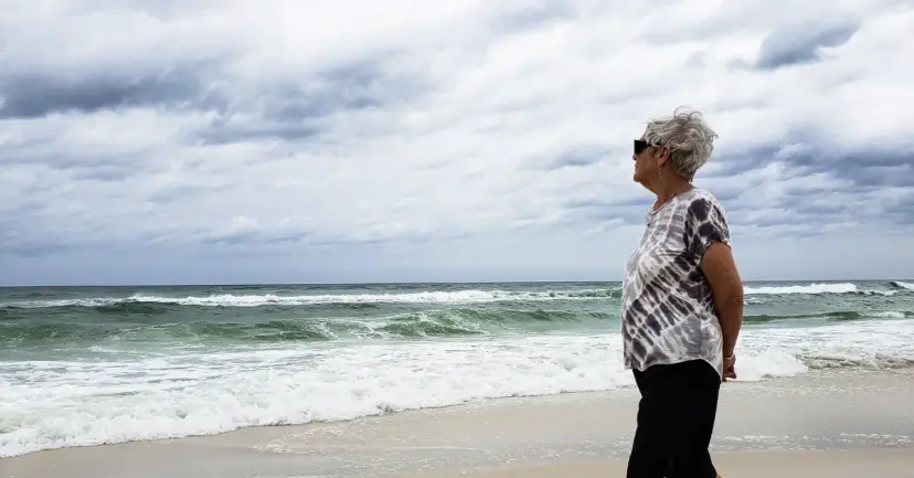 An older woman in sunglasses stands on a beach looking out at breaking waves under clouds.