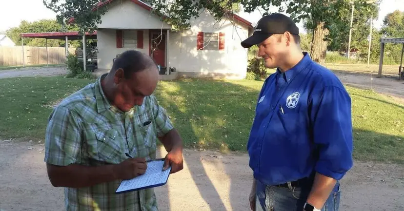 A man signing a petition on a clipboard while a man in a blue TNM shirt watches outside a house.