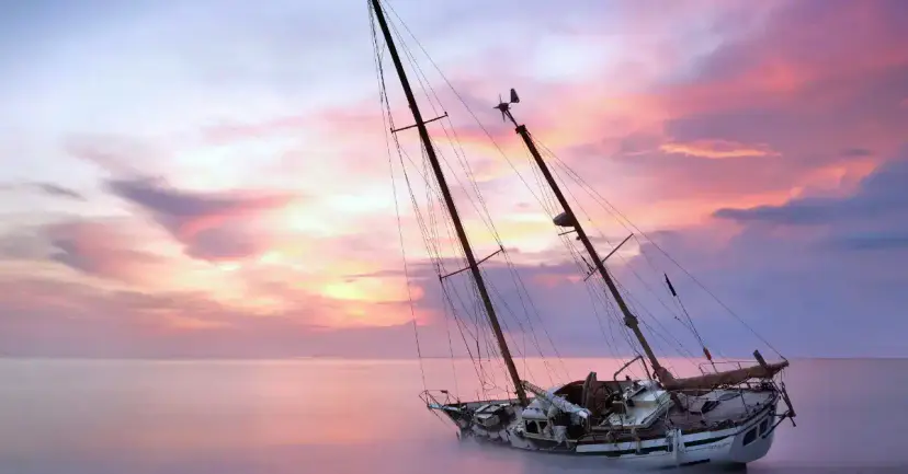 A sailboat lying tilted and aground in shallow water under a pink sunset sky.
