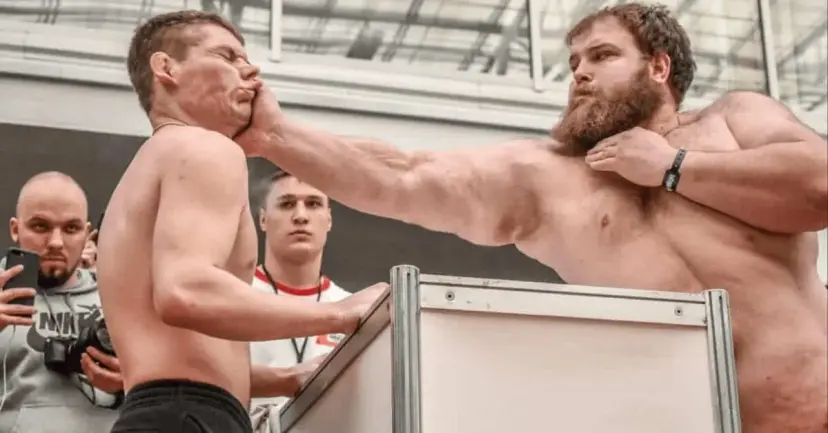 Two shirtless men at a slap competition, one striking the other across the face as spectators watch.