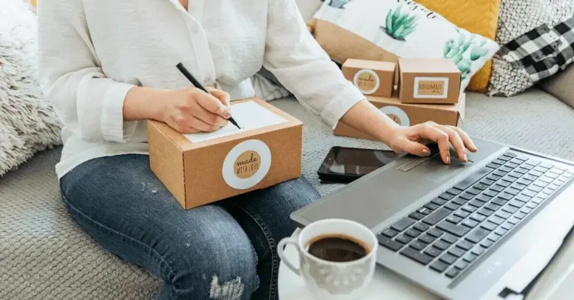 A woman labels a shipping box while working on a laptop at home.