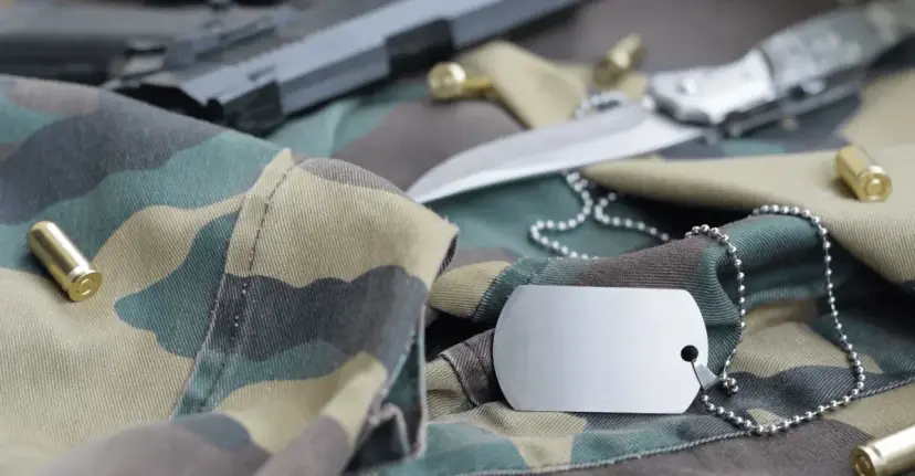 Military dog tags, a knife, a handgun and brass cartridges lying on camouflage fabric.
