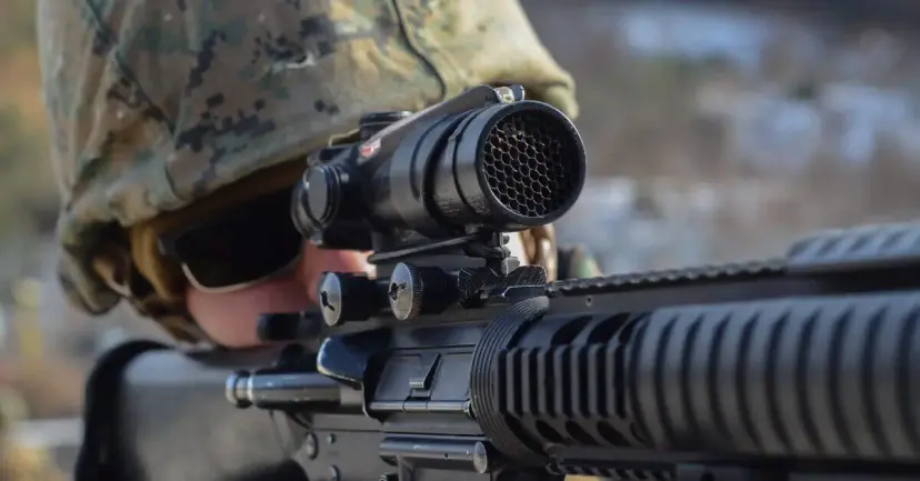 A soldier in camouflage aims a rifle fitted with a mounted optical sight.