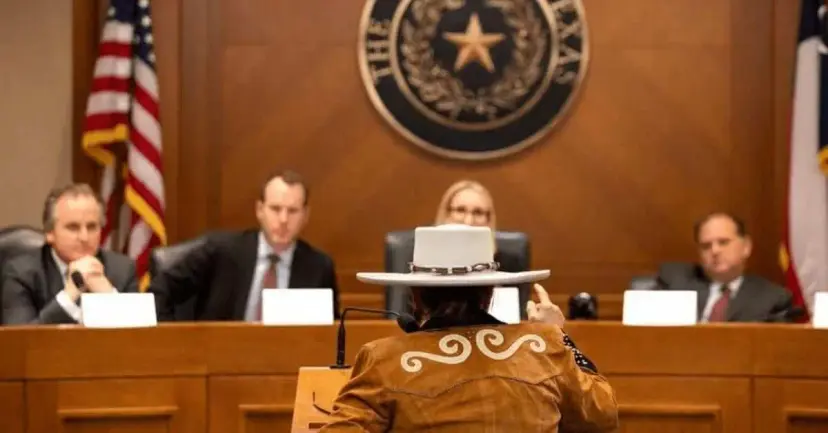 A man in a white cowboy hat and fringed jacket testifying before a panel of officials beneath the Texas seal.