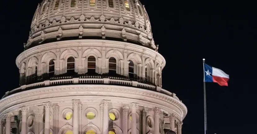 The Texas Capitol dome lit up at night with a Texas flag flying beside it.