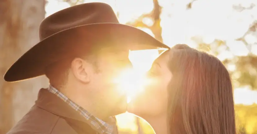 A man in a cowboy hat kisses a woman outdoors with the sun shining between them.