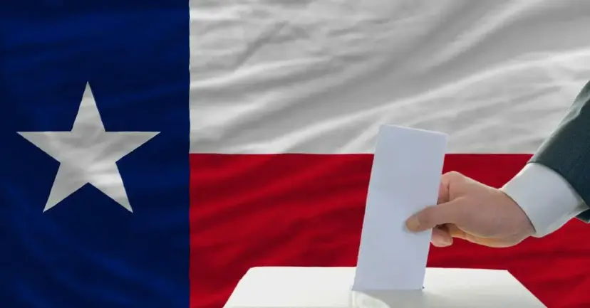 A hand dropping a paper ballot into a box in front of a large Texas flag.