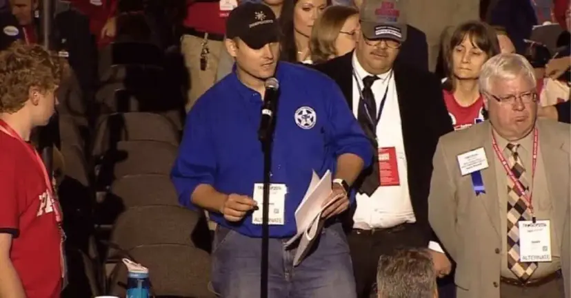 A man in a blue TNM shirt speaks at a floor microphone at a crowded political convention.