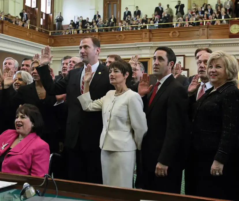 Lawmakers raising their right hands to take an oath in a legislative chamber, with a crowded gallery above.