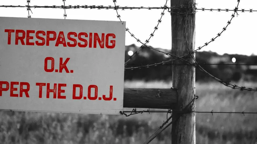 Sign on a barbed wire fence reading 'Trespassing O.K. per the D.O.J.' in red letters.