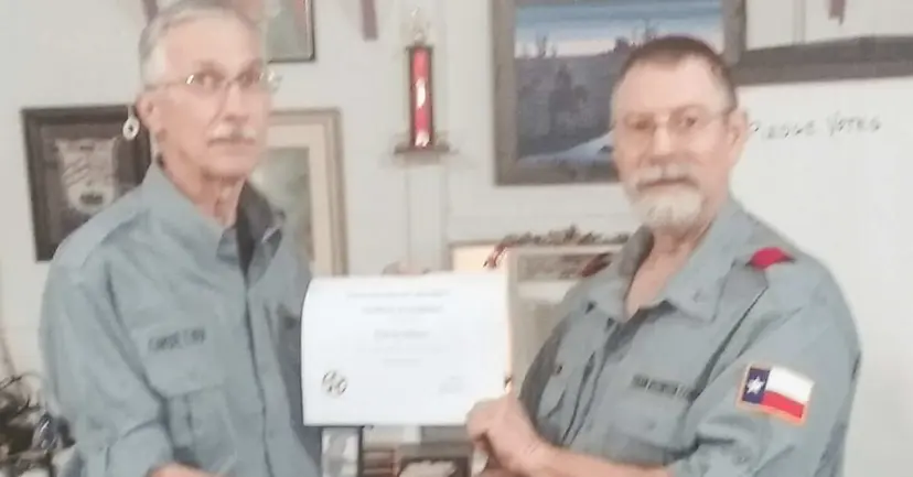 Two older men in gray uniform shirts holding a certificate between them in a room with framed pictures.