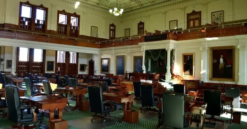 Empty Texas Senate chamber with wooden desks, leather chairs, green carpet and portraits on the walls.