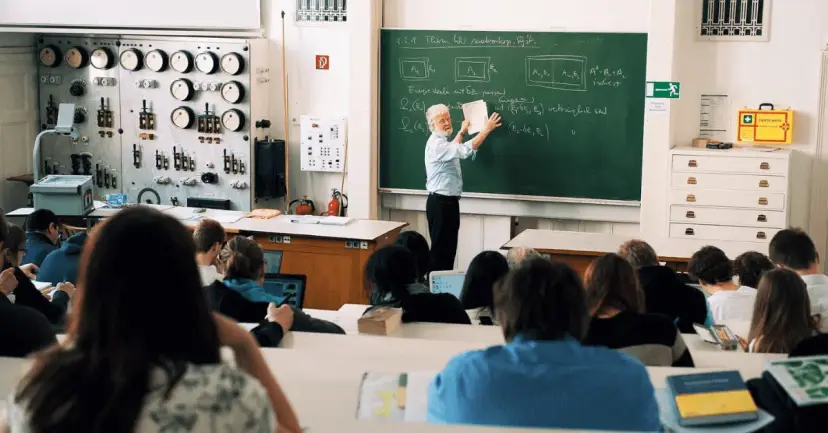 A professor writes on a green chalkboard while university students listen in a lecture hall.