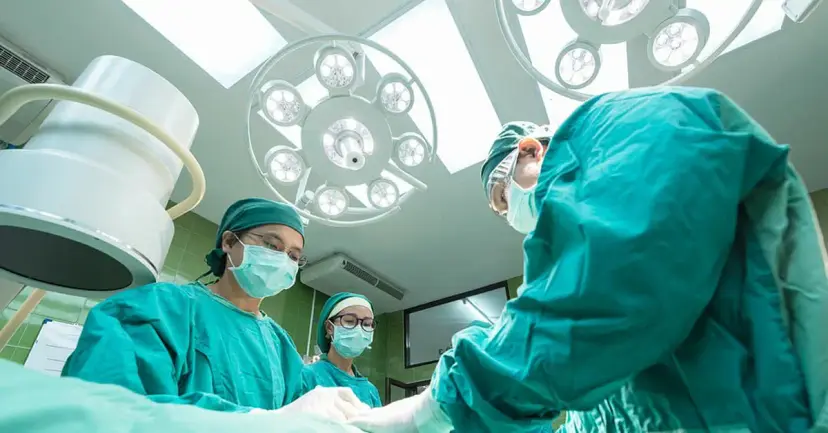 Surgeons in green scrubs, caps and masks working under bright lights in an operating room.