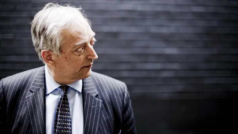 Gray-haired man in a pinstriped suit and patterned tie glancing to one side against a dark wall.