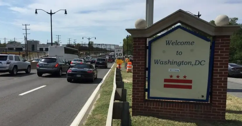 Traffic on a busy road passing a brick sign reading "Welcome to Washington, DC" with the city flag.