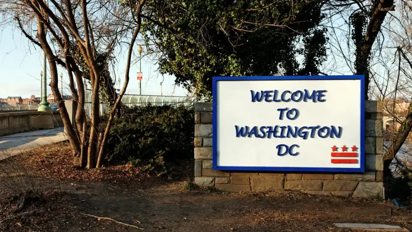 Sign reading 'Welcome to Washington DC' on a stone base beside bare trees and a walking path.