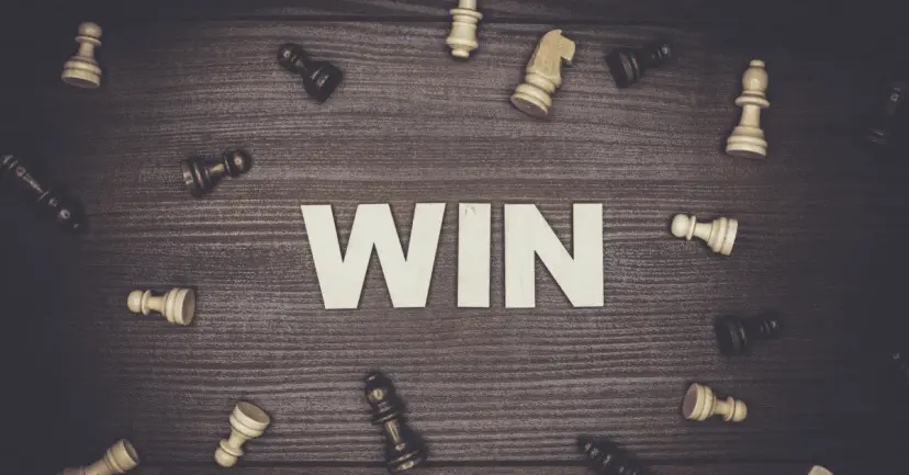 Wooden letters spelling 'WIN' in the center of scattered chess pieces on a dark wood surface.