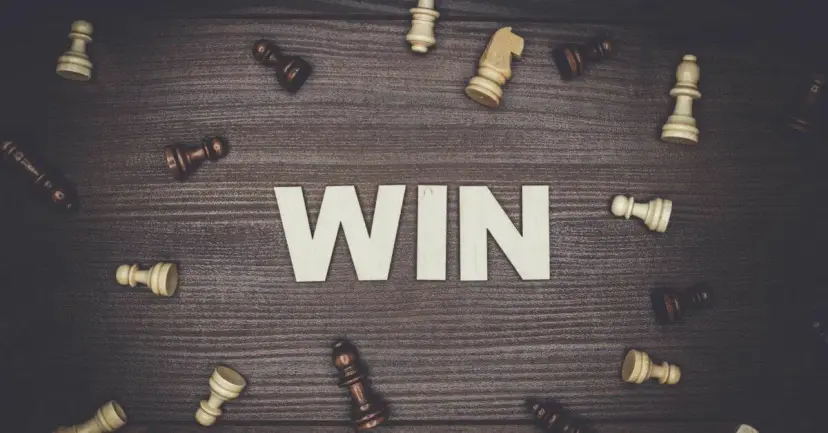 The word 'WIN' in wooden letters surrounded by scattered chess pieces on a dark wood table.