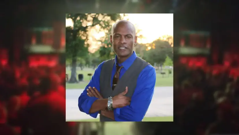 Claver Kamau-Imani standing with arms crossed in a blue shirt and gray vest, framed over a blurred red crowd.