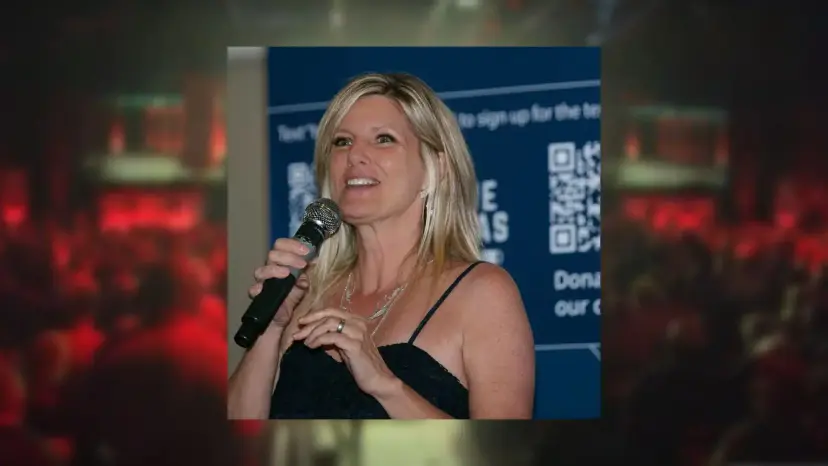 Julie McCarty speaks into a microphone in front of a blue sign-up banner, set over a blurred red background.