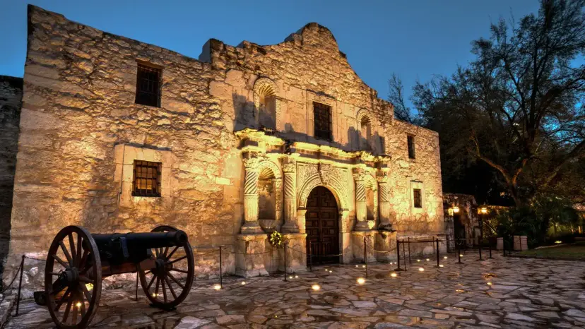 The Alamo chapel lit at dusk, with an old cannon on the stone plaza in front.