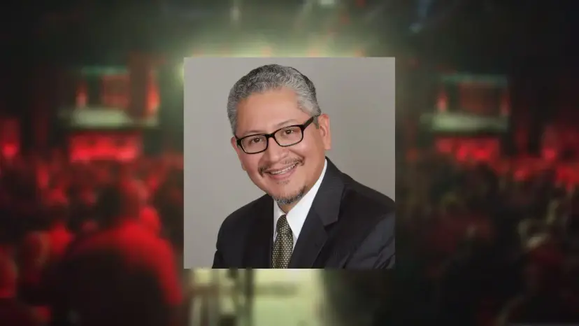 Headshot of Carlos Lopez in glasses and a suit, set over a blurred red concert hall background.