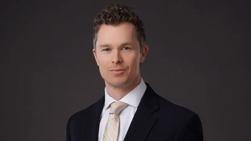Paul Davis, a man with short curly hair in a dark suit and striped tie, on a gray background.