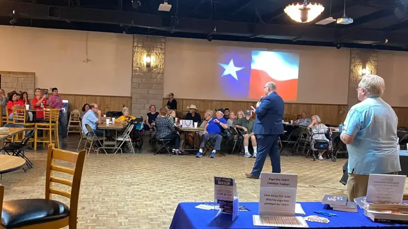 A speaker in a suit addresses a seated crowd in a hall with a Texas flag projected on the wall.