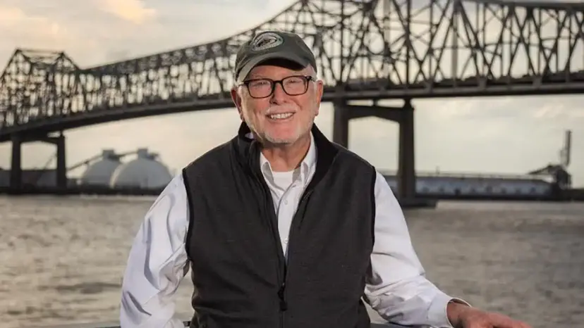 Richard Fossey in a cap and vest smiles on a riverbank with a steel bridge behind him.