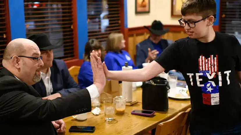 At a restaurant table, a boy in a TEXIT shirt high-fives a bald man in a suit.