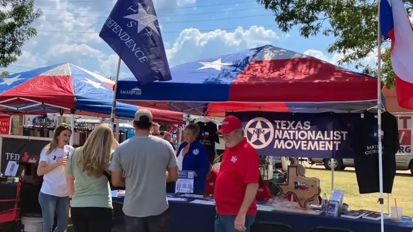 Texas Nationalist Movement booth under Texas flag canopies at an outdoor festival, with volunteers talking to visitors.