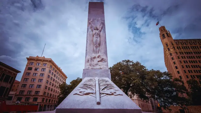 The Alamo Cenotaph in San Antonio seen from below, a carved stone monument against a cloudy sky between downtown buildings.