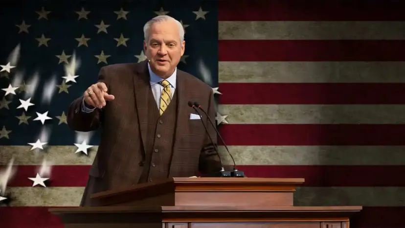 Gray-haired man in a brown three-piece suit speaks at a wooden podium in front of an American flag.