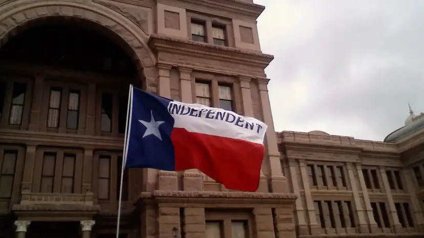 Texas flag printed with the word 'Independent' flies in front of the red granite Texas Capitol.