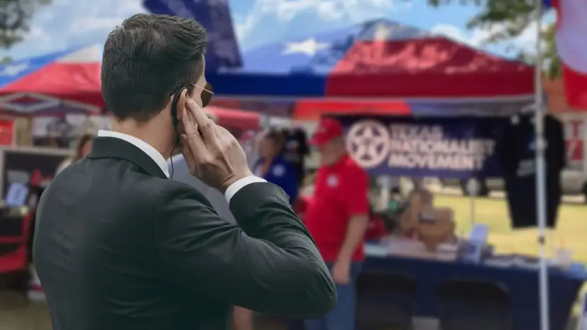 A security agent in a dark suit with an earpiece watching a Texas Nationalist Movement booth at an outdoor event.