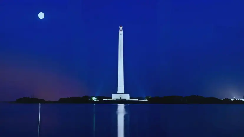 San Jacinto Monument lit up at night under a full moon, reflected in still water.