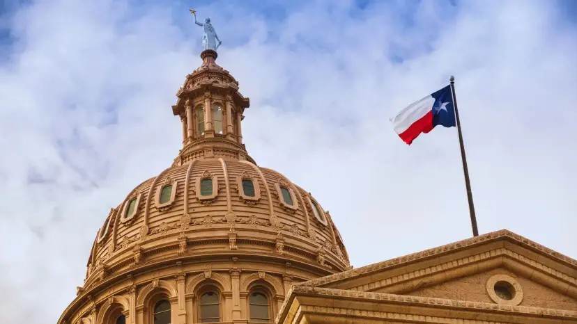 The Texas Capitol dome in Austin with a Texas flag flying against a cloudy sky.