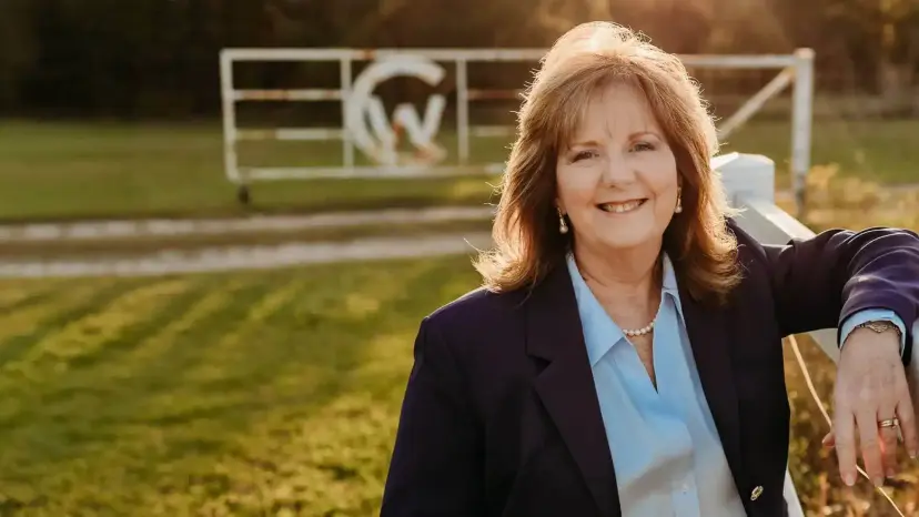 Paulette Carson smiling in a navy blazer, leaning on a fence in front of a ranch gate.