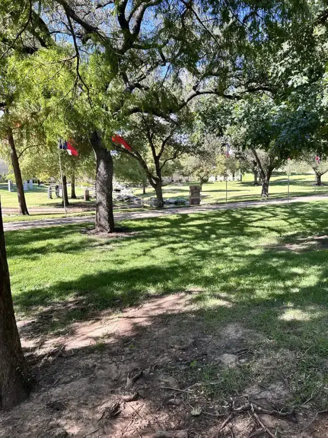 Shaded cemetery grounds under oak trees, with Texas flags flying among the headstones in the distance.