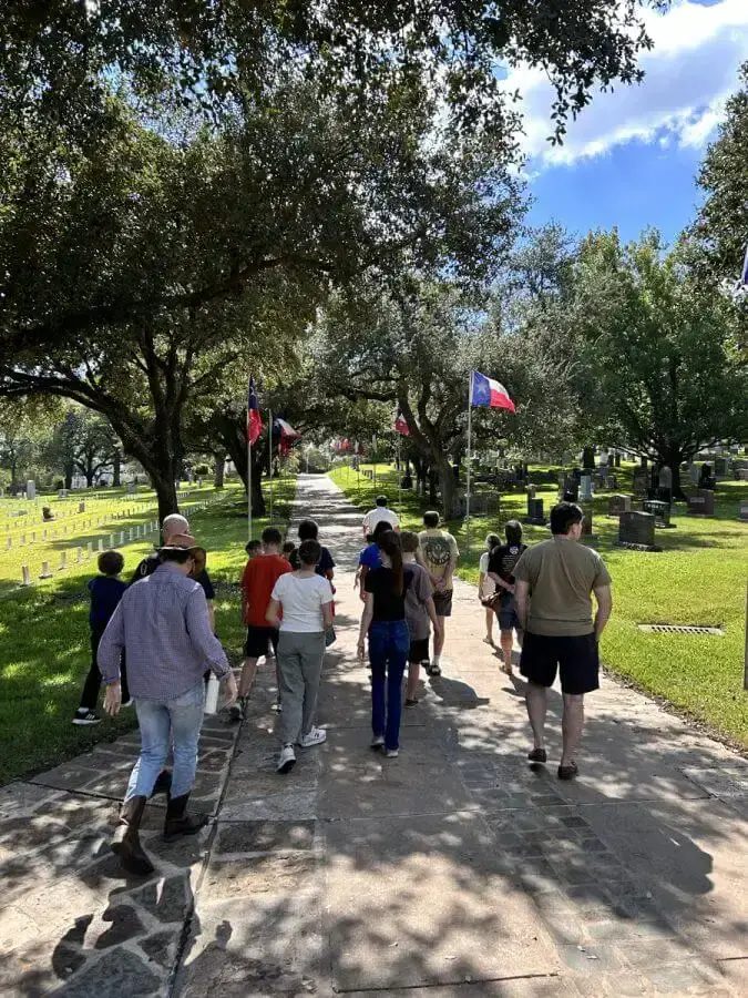 Group of children and adults walk down a shaded cemetery path lined with Texas flags.
