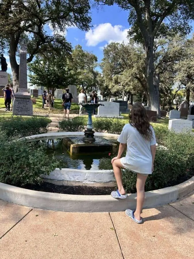 Girl rests a foot on the edge of a cemetery fountain as a tour group gathers near monuments.