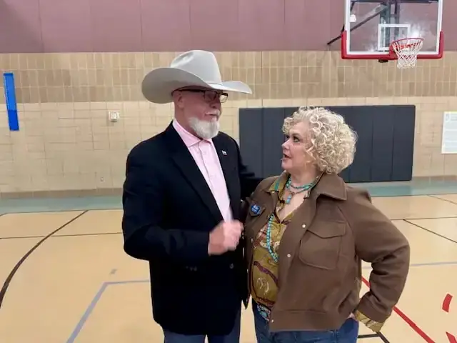 A man in a white cowboy hat talks with a woman in a brown jacket in a school gym.